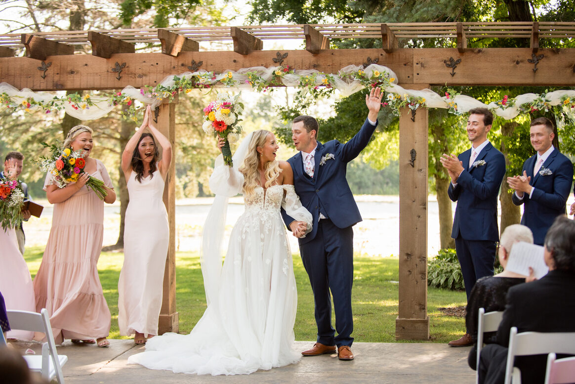 A bride and groom celebrate with raised arms beneath the floral pergola at their outdoor ceremony at The Chart House