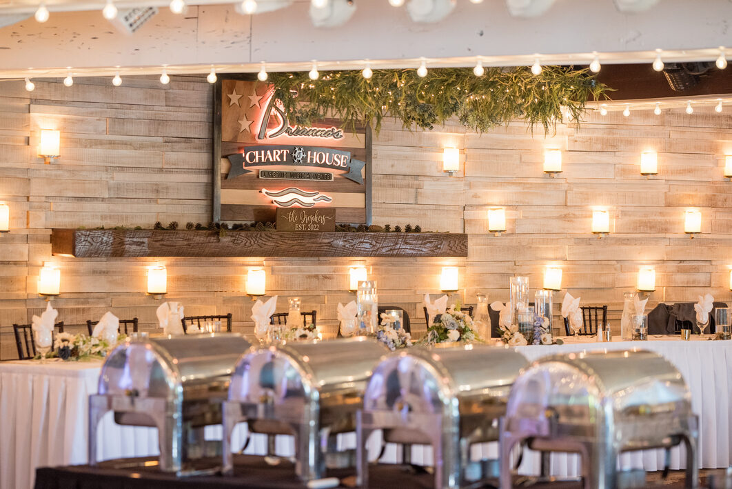 A buffet of chafing dishes set under string lights in front of the Chart House sign for a corporate gathering