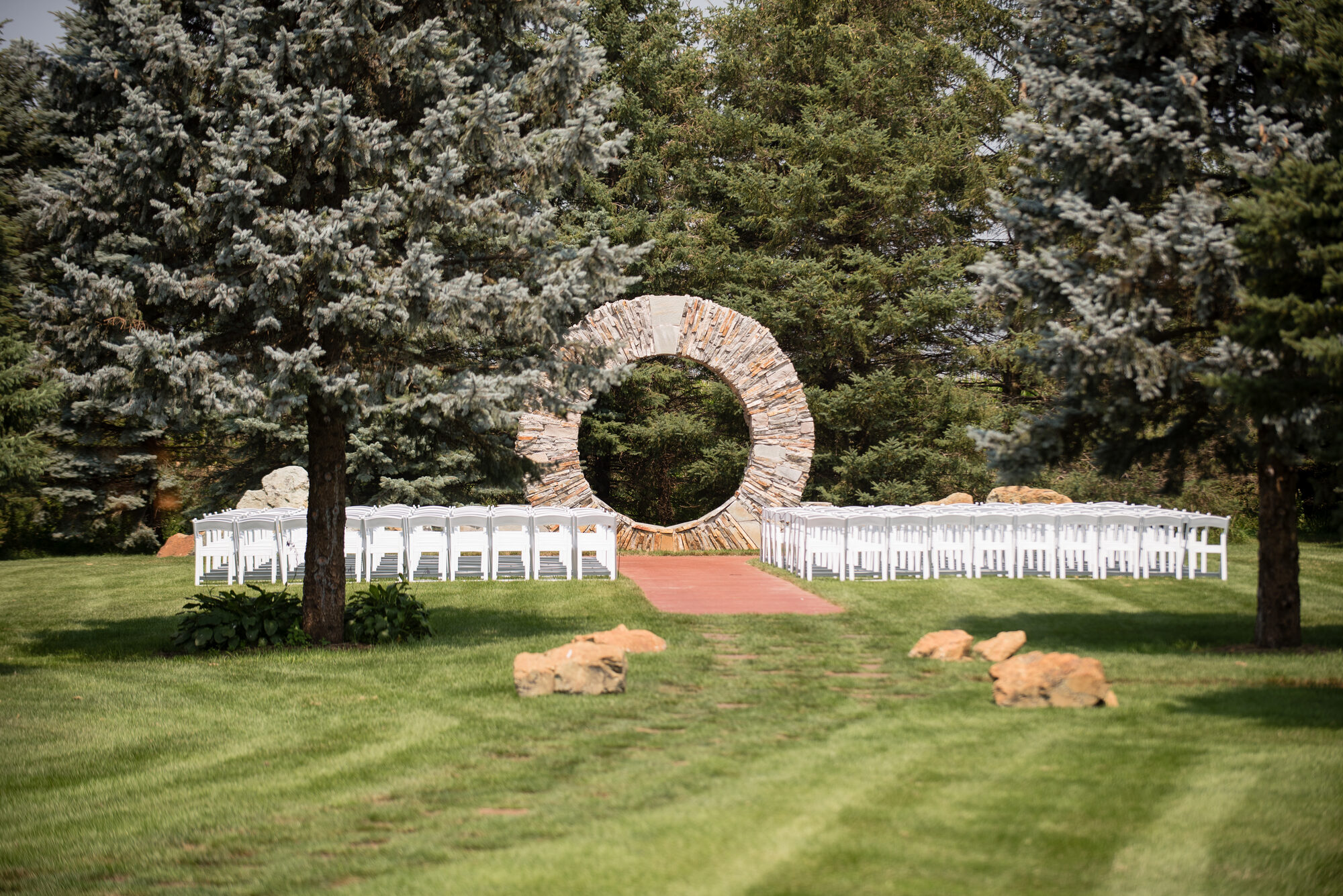 Outdoor garden ceremony with a stone moon-gate arch and rows of white chairs framed by evergreens at Glenhaven Events, Farmington MN