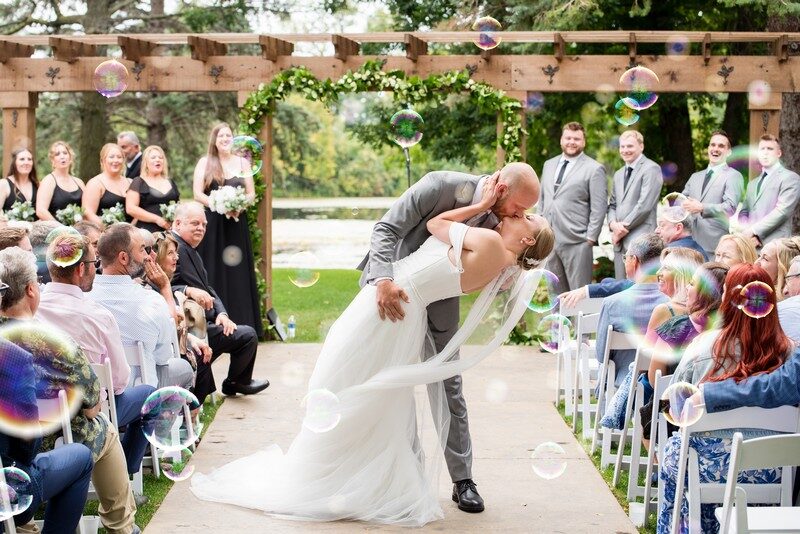 Newlyweds kiss as guests blow bubbles down the aisle at an outdoor ceremony at The Chart House, Lakeville MN