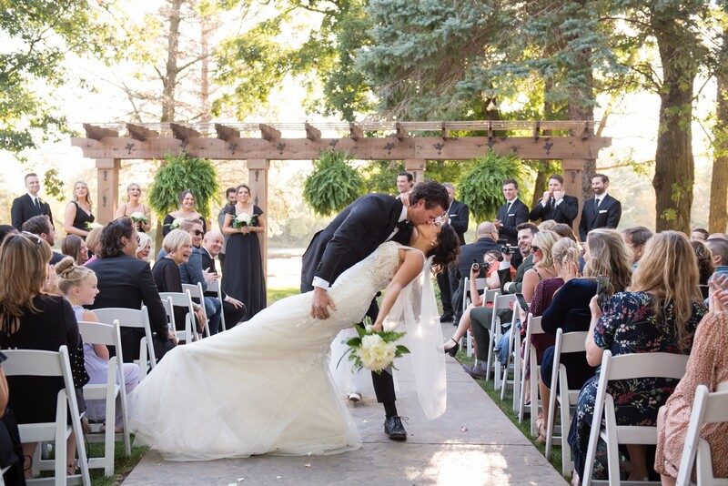 Bride and groom share their first kiss beneath the greenery-draped pergola during an outdoor garden ceremony at The Chart House, Lakeville MN