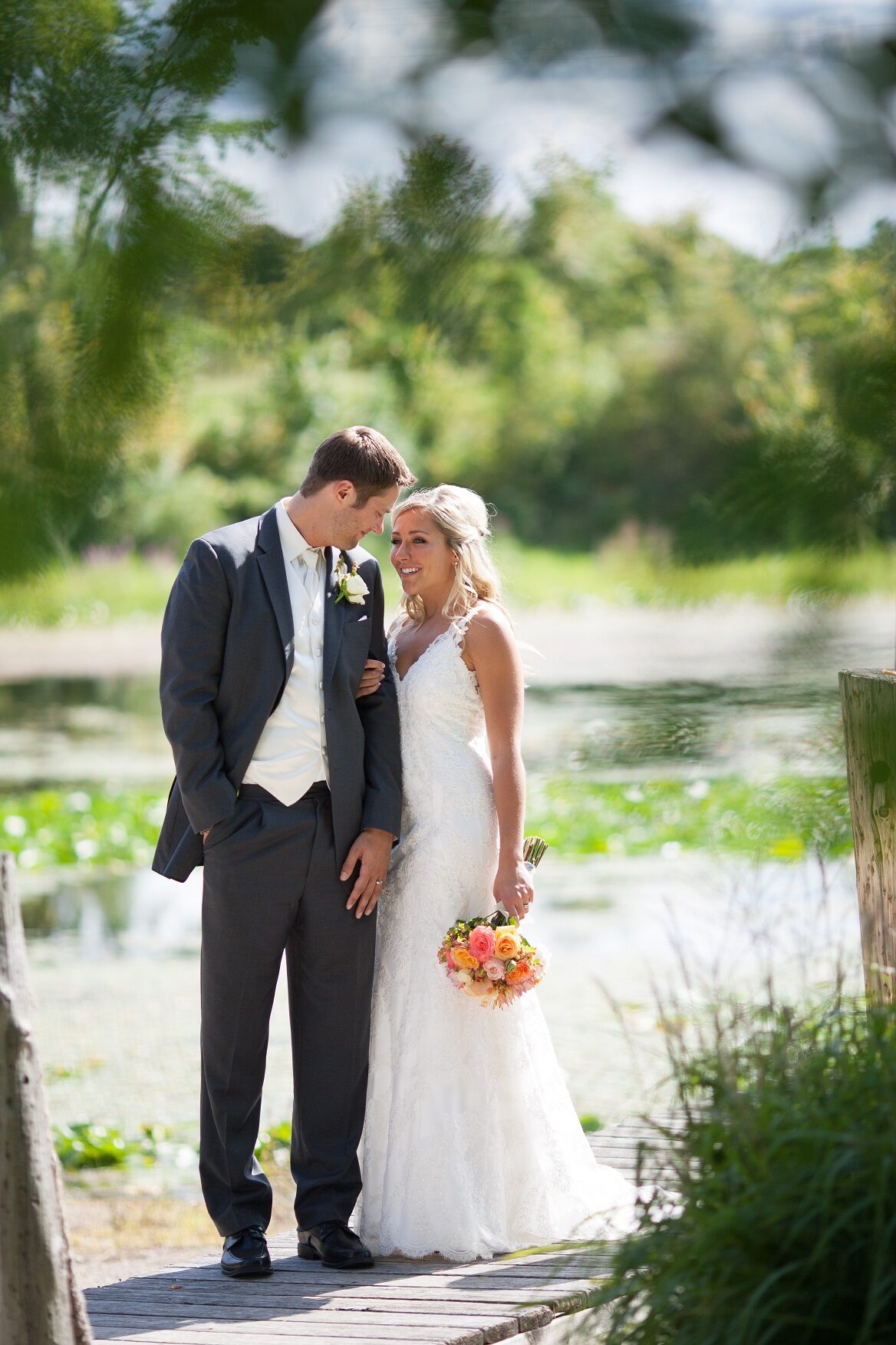 Bride and groom embrace on the dock with the lake and summer greenery behind them at The Chart House, Lakeville MN