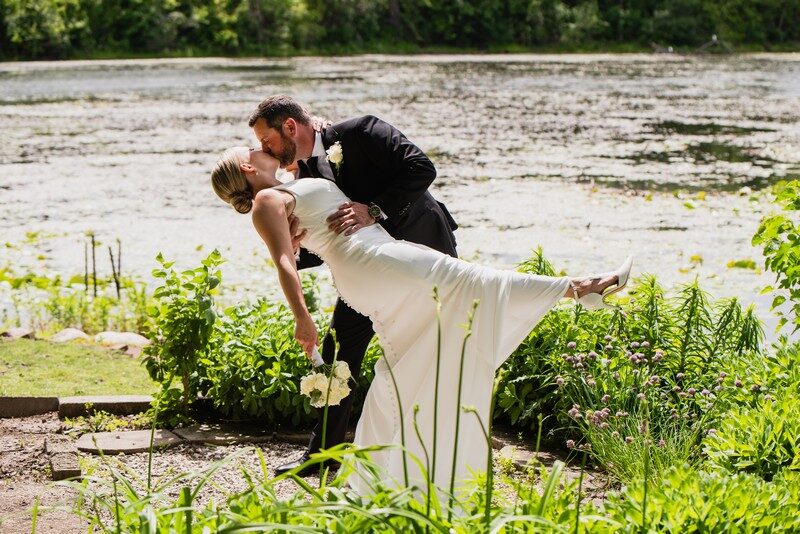 Groom dips his bride for a kiss at the water's edge amid lush summer greenery at The Chart House, Lakeville MN