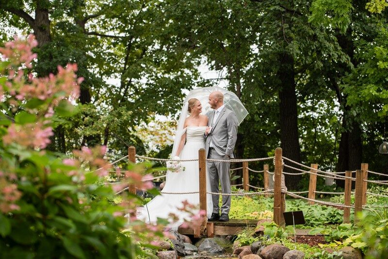Bride and groom share a clear umbrella on the garden bridge among blooming hydrangeas at The Chart House, Lakeville MN