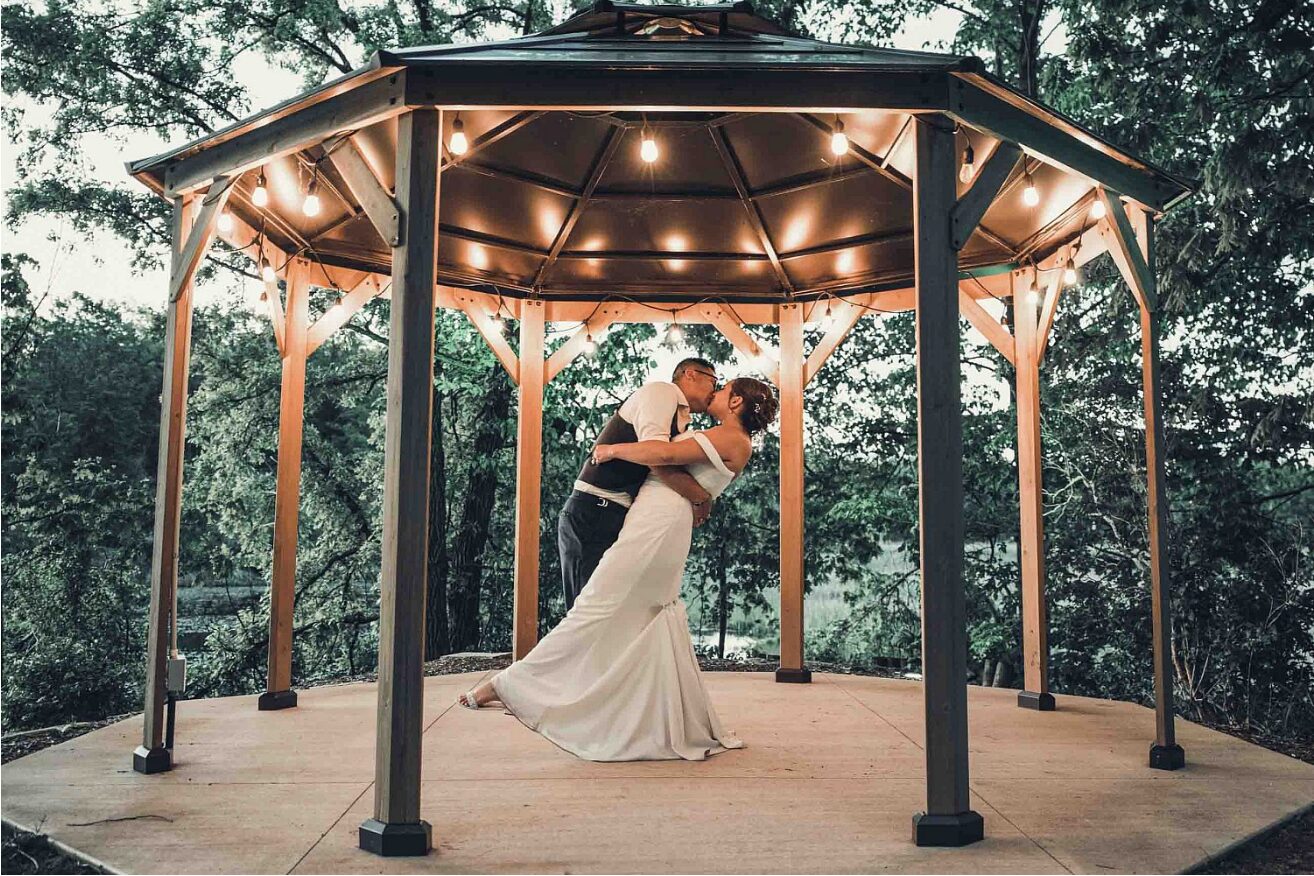 Bride and groom kiss beneath the string-lit lakeside gazebo at dusk at The Chart House, Lakeville MN