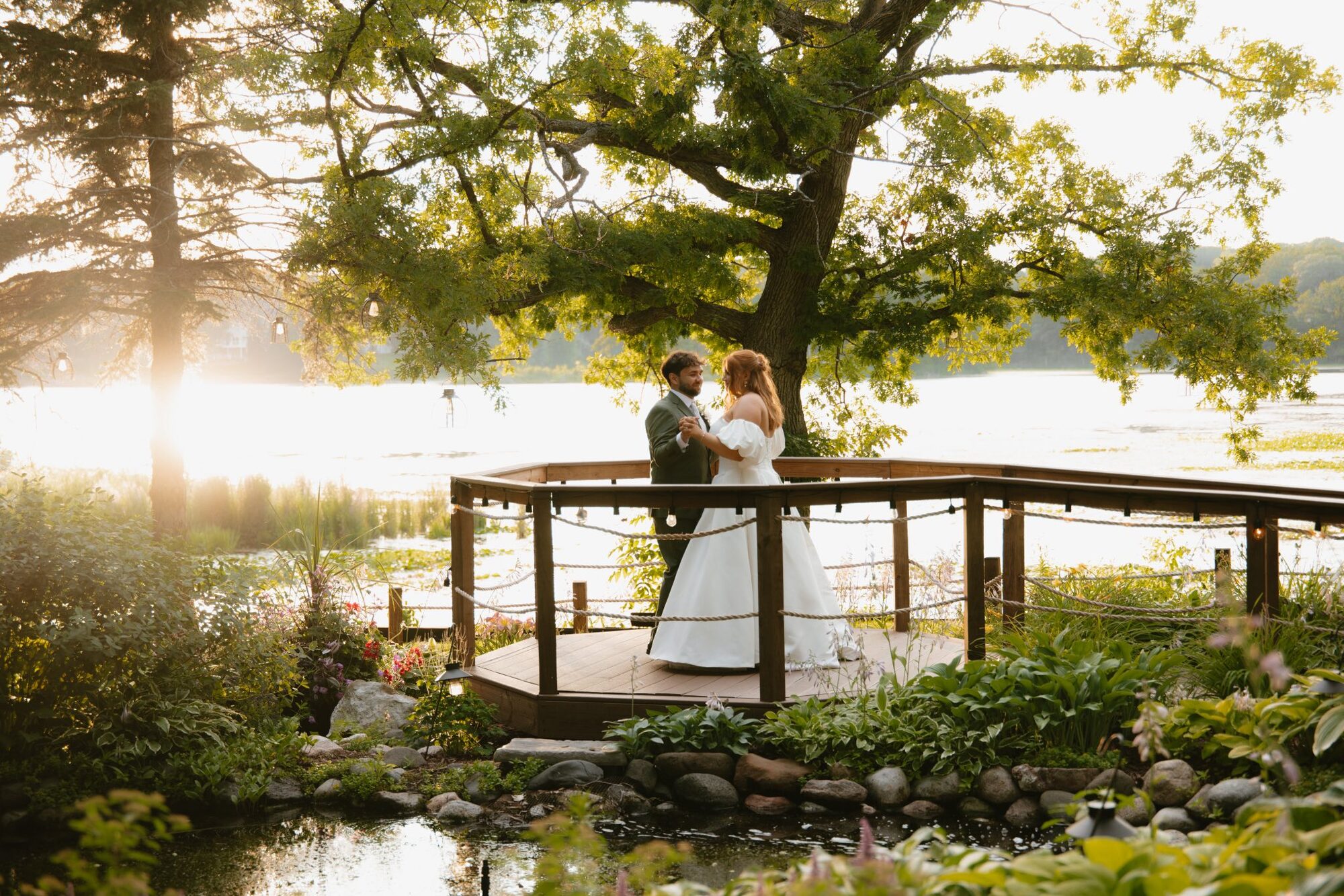 Bride and groom dance on the lakeside deck under a sprawling oak tree at golden hour at The Chart House, Lakeville MN