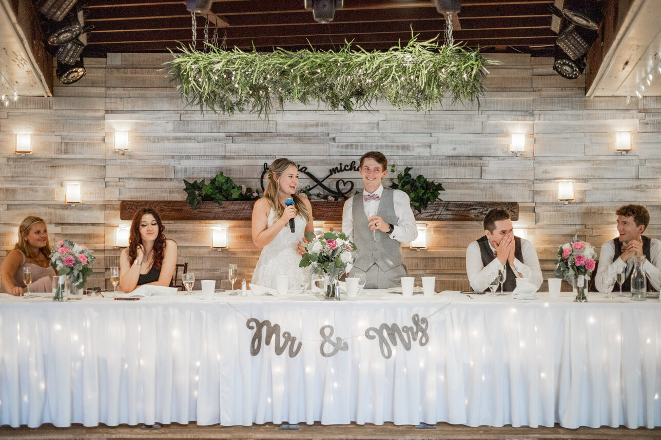 Bride and groom give a speech at the skirted head table beneath a greenery garland and 'Mr & Mrs' banner at The Chart House, Lakeville MN
