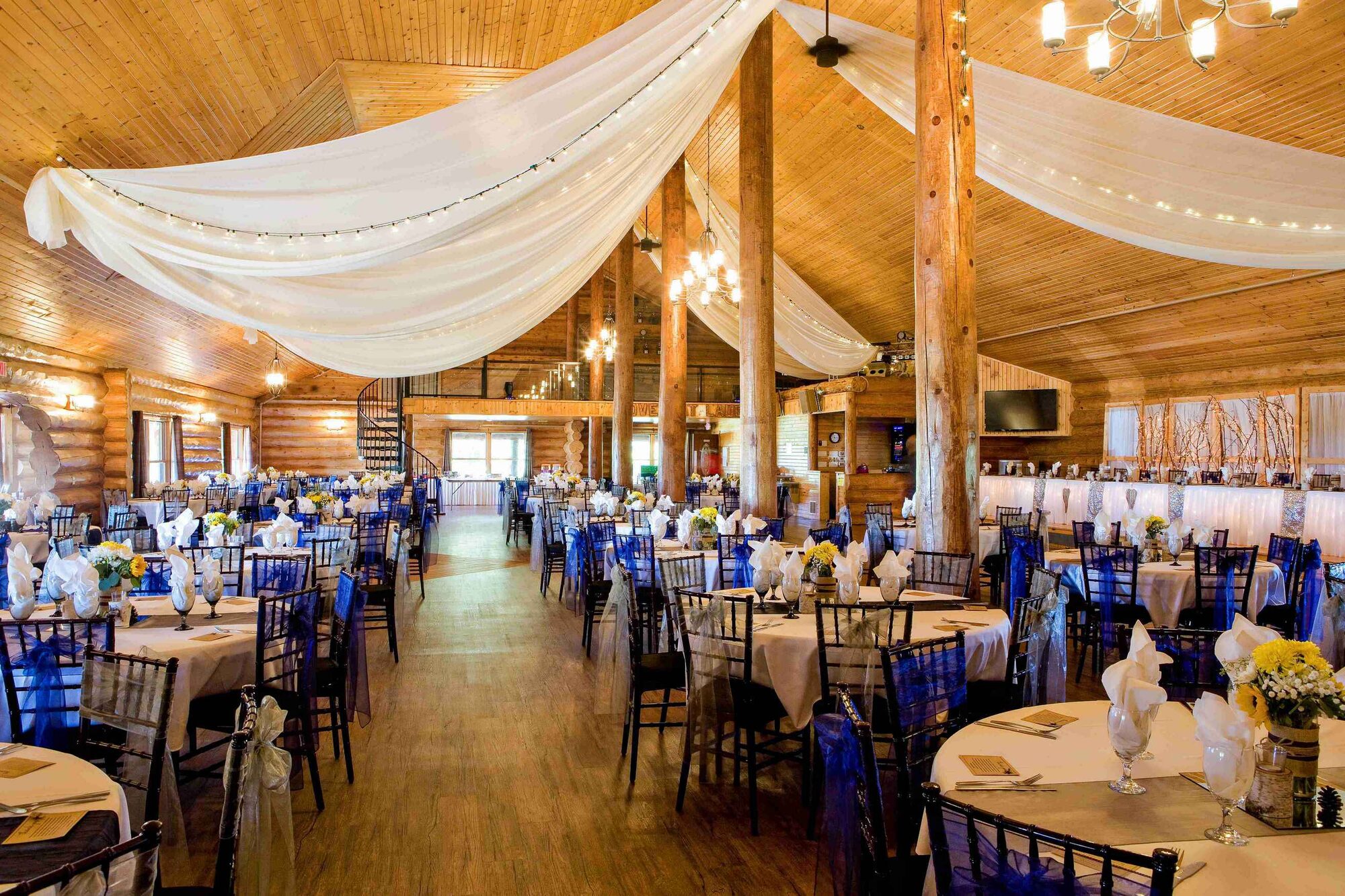Log lodge ballroom with white ceiling drapery, string lights, and blue-sashed chairs set for a reception at Glenhaven Events, Farmington MN
