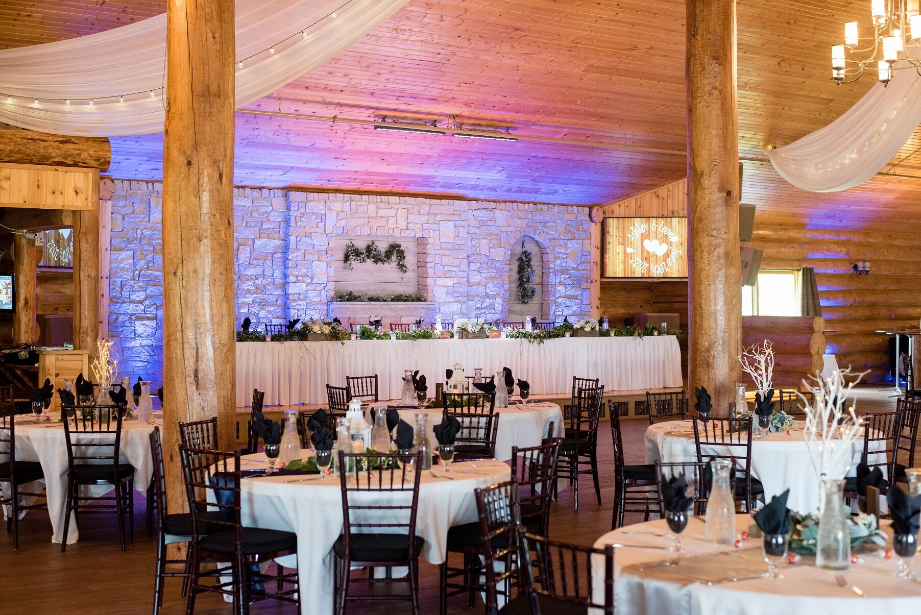 Log-and-stone ballroom set with round tables under purple and blue uplighting beside the stone fireplace at Glenhaven Events, Farmington MN