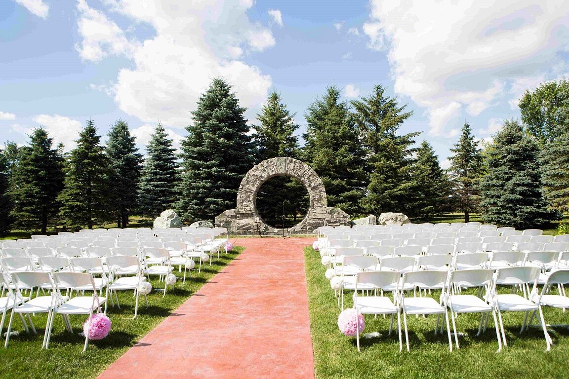 Outdoor ceremony site with a stone moon-gate arch framed by evergreens and rows of white chairs at Glenhaven Events, Farmington MN