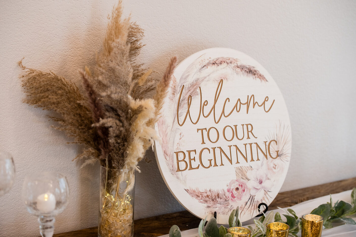 A round 'Welcome to our beginning' wood sign with pampas grass, florals, and candles on a reception table