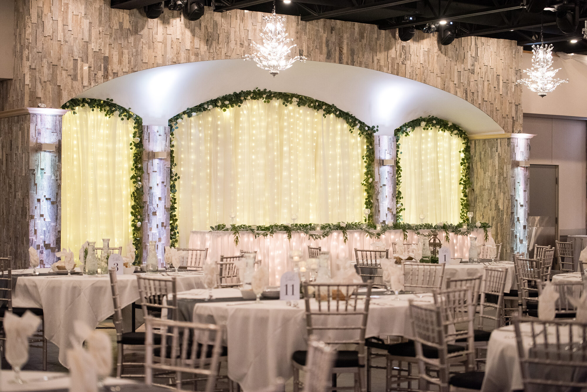 Royal Cliff ballroom set for a reception with silver Chiavari chairs, round tables, a fairy-light draped backdrop with greenery, and chandeliers, Eagan MN