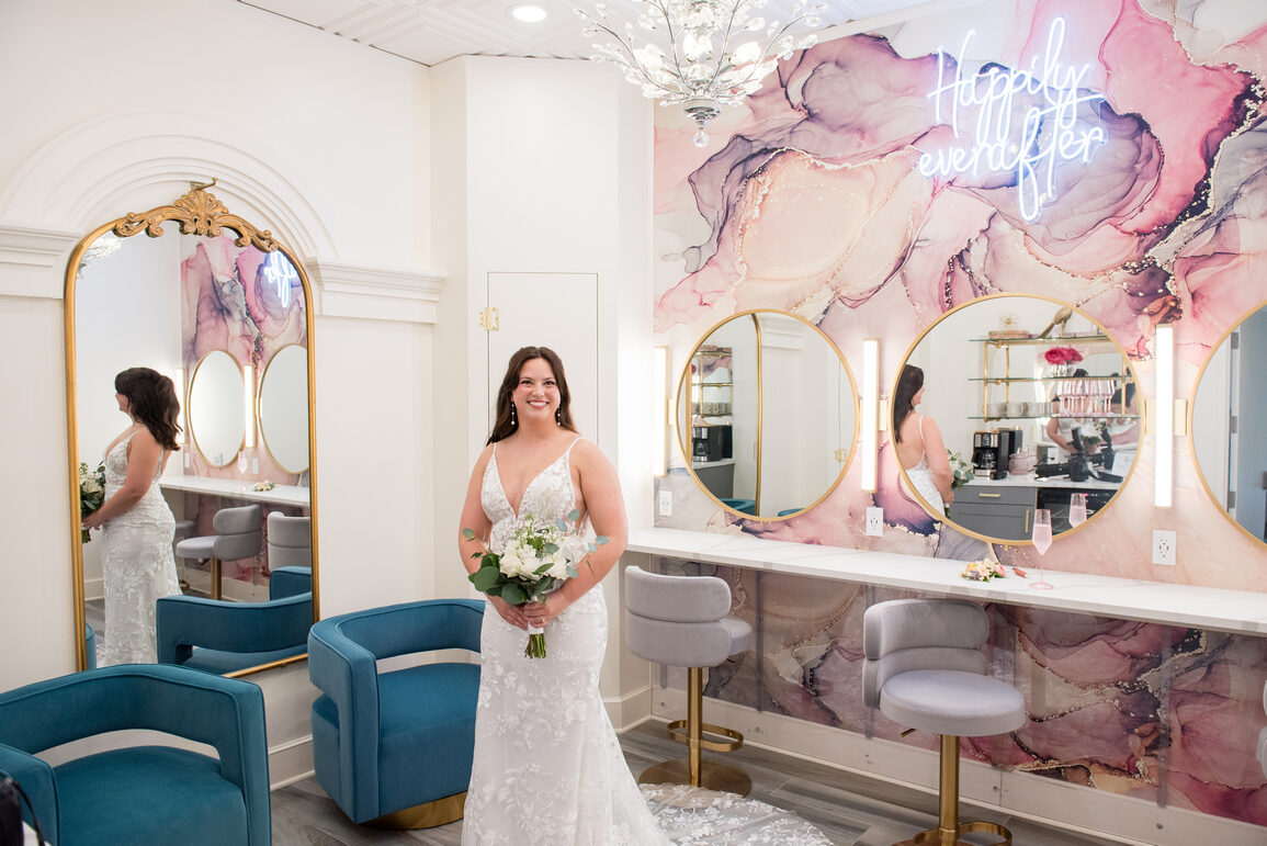 Bride in a lace gown holding a white bouquet beside the Happily Ever After neon and pink mural in the bridal salon at Royal Cliff, Eagan MN