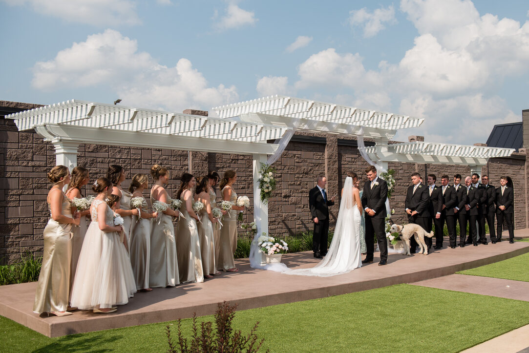 Bride and groom exchanging vows under a white pergola flanked by the wedding party, with their dog, in the stone-walled garden at Royal Cliff, Eagan MN