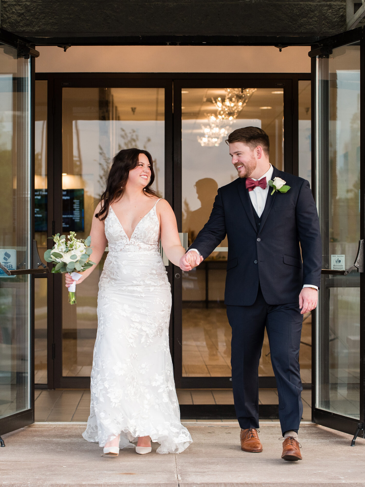 Bride and groom walk hand in hand out the glass front entrance beneath a chandelier at Royal Cliff, Eagan MN