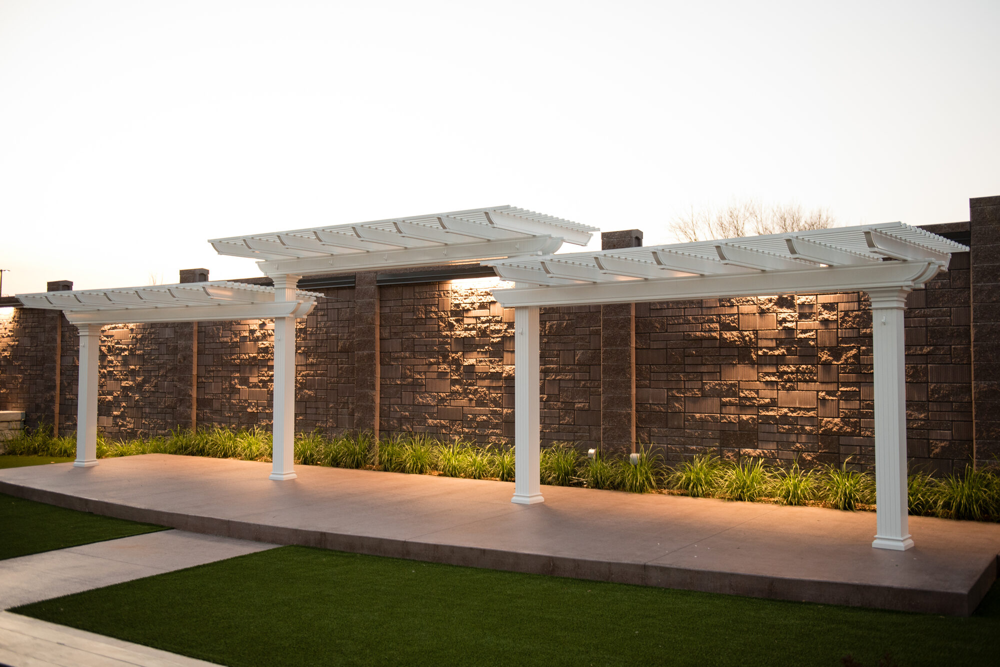 White pergolas over the stone walkway in the outdoor ceremony garden against the textured stone wall at golden hour at Royal Cliff, Eagan MN