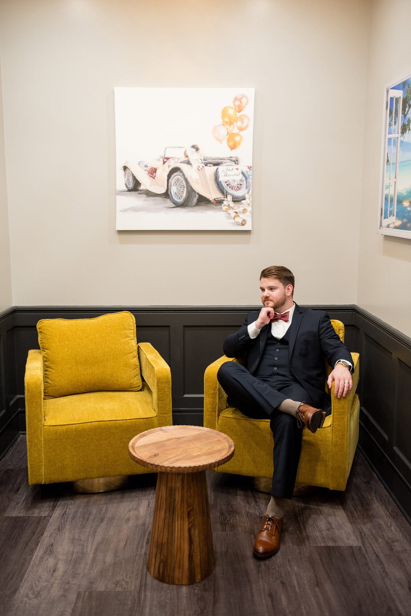 Groom relaxing in a mustard-yellow armchair beside a wood side table and a Just Married canvas in the groom's room at Royal Cliff, Eagan MN