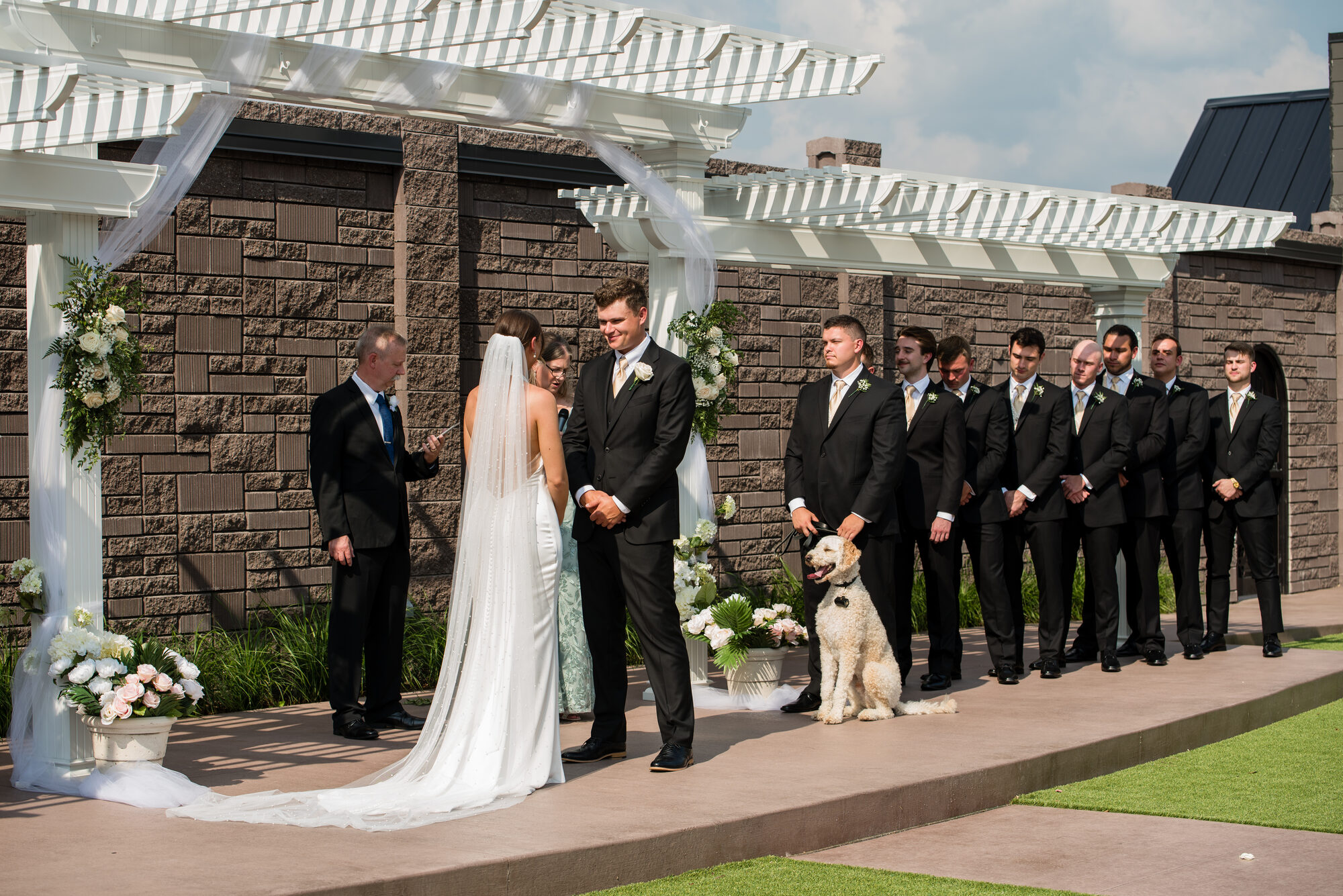Outdoor garden ceremony beneath the white pergola at Royal Cliff — bride and groom at the altar with their wedding party and dog, Eagan MN