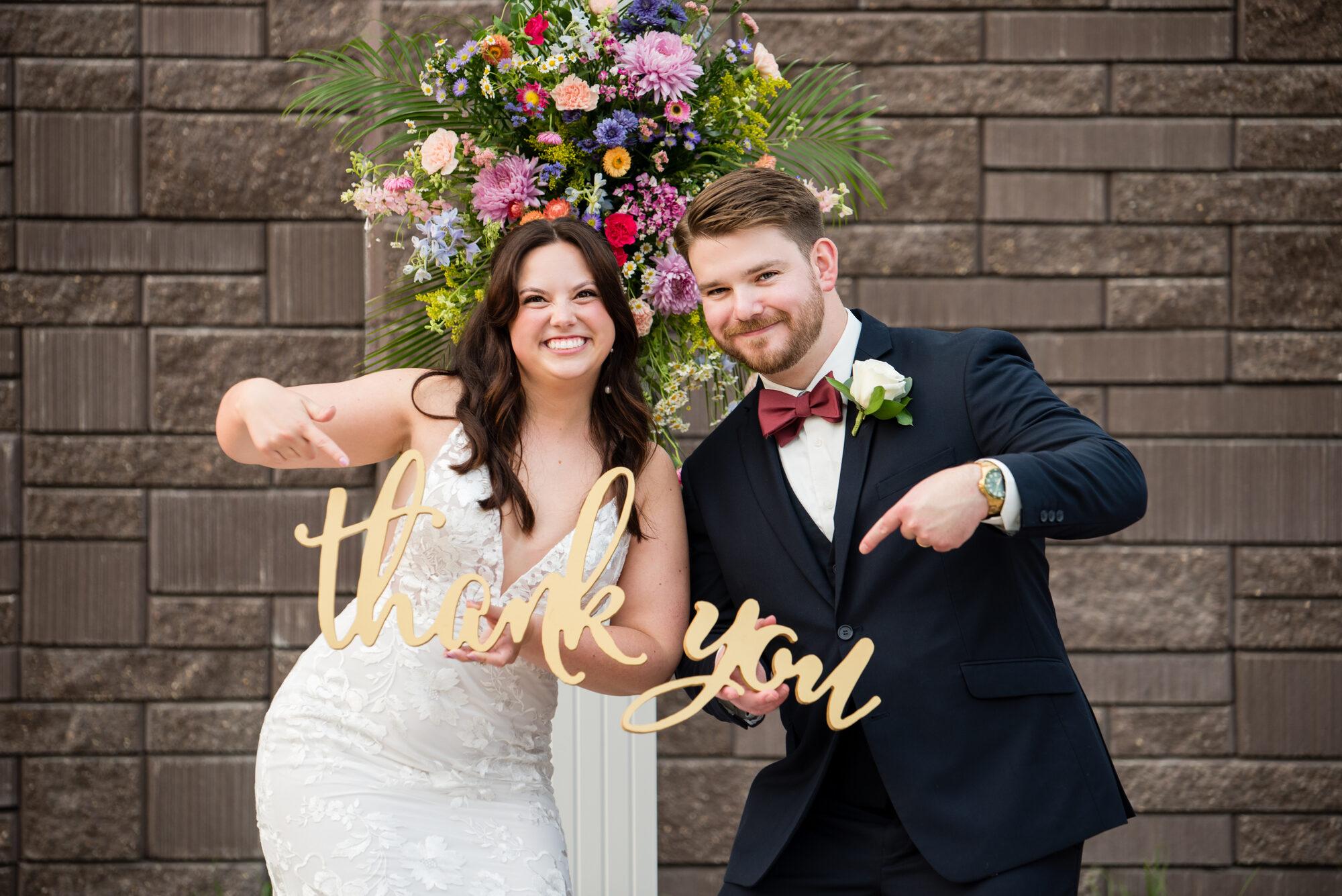 Bride and groom holding a gold script thank-you sign in front of a colorful floral arrangement against the stone wall at Royal Cliff, Eagan MN