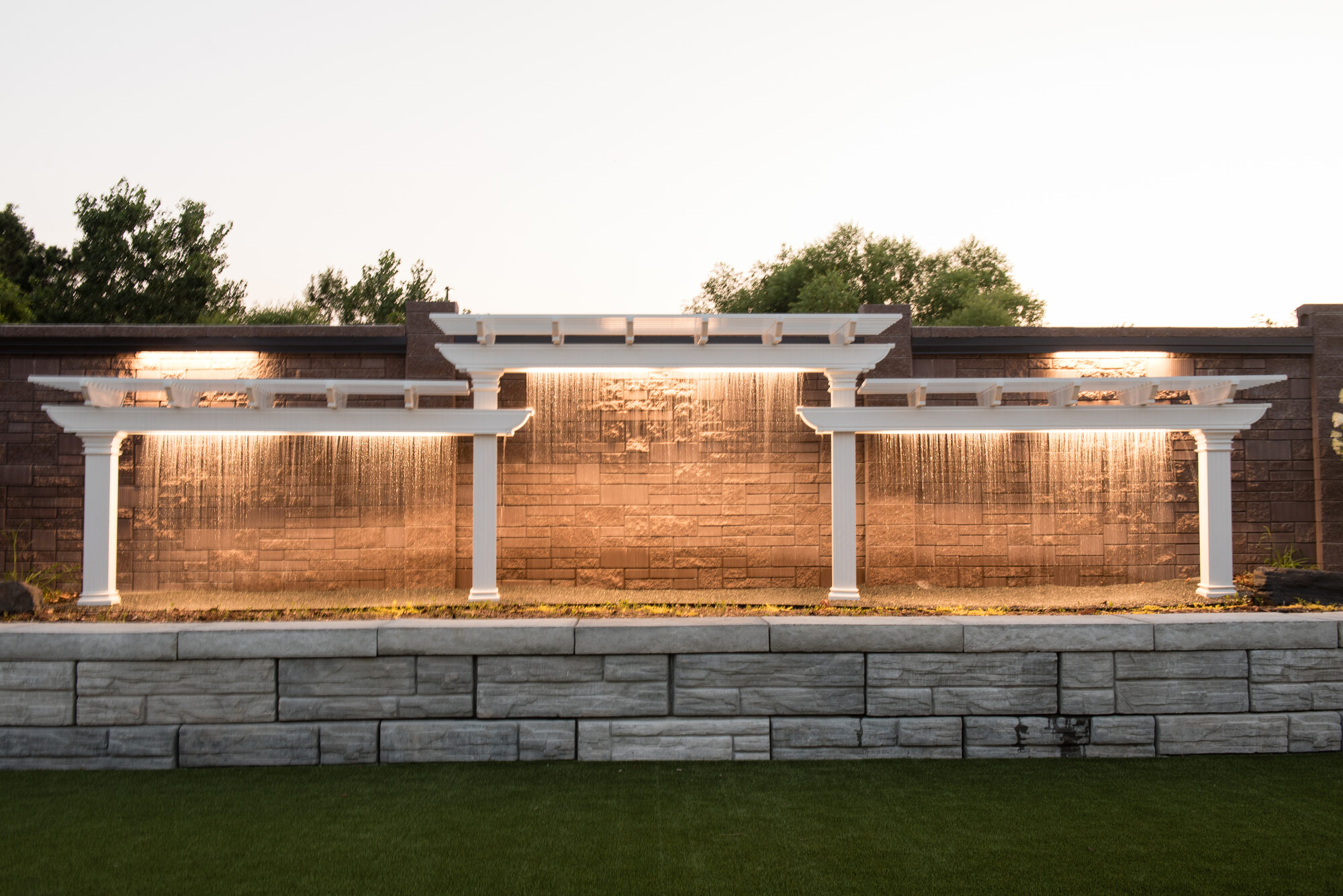 Illuminated water curtain cascading down the stone wall between white pergolas at golden hour in the outdoor garden at Royal Cliff, Eagan MN